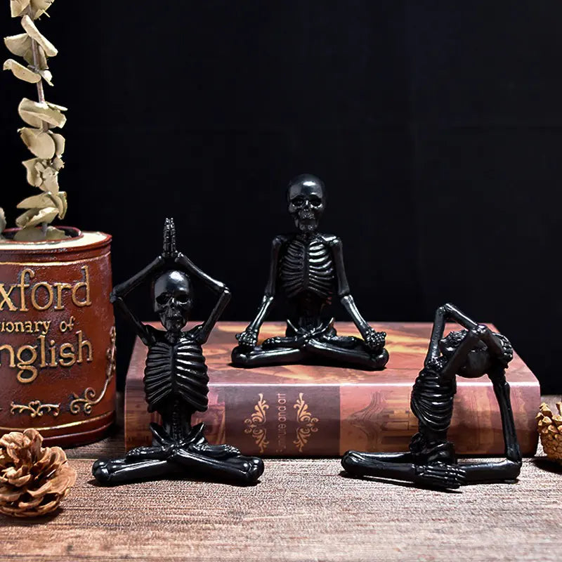 Three black skeleton figurines in yoga poses on a wooden surface with a book and decorative items.