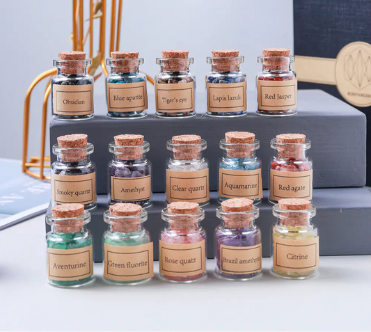 Collection of small glass jars with cork lids containing different types of gemstones, labeled with names, on a white surface.