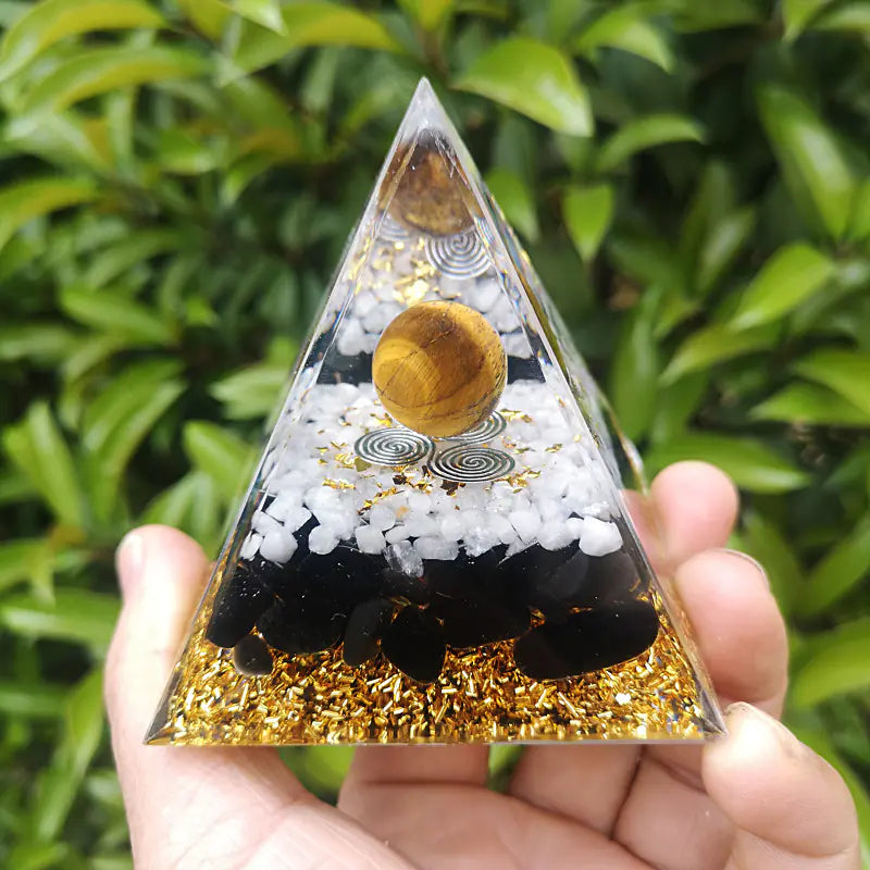 Pyramid-shaped object with black stones, white pebbles, and gold glitter held in front of green foliage.