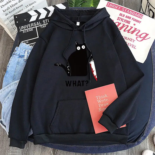 Black hoodie with a cat graphic and 'WHAT?' text on a wooden surface with books and jeans.