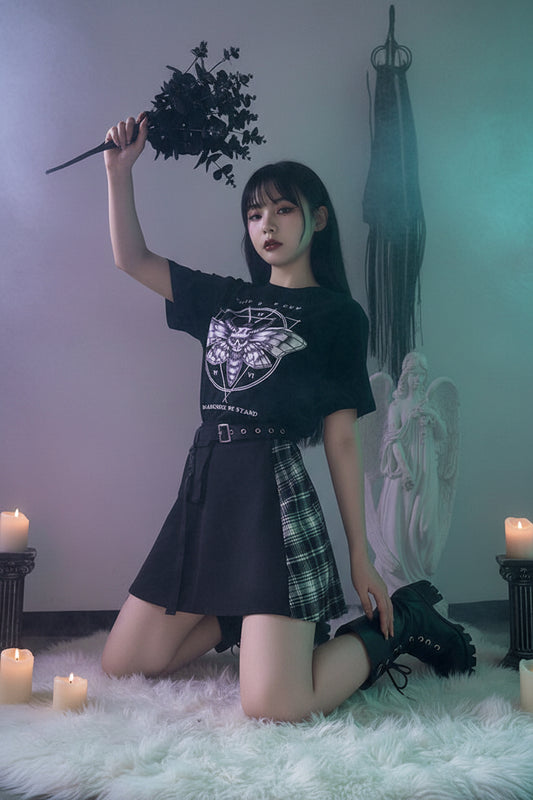 Person wearing a black t-shirt with a moth design and text, holding dark foliage, standing in front of a statue.