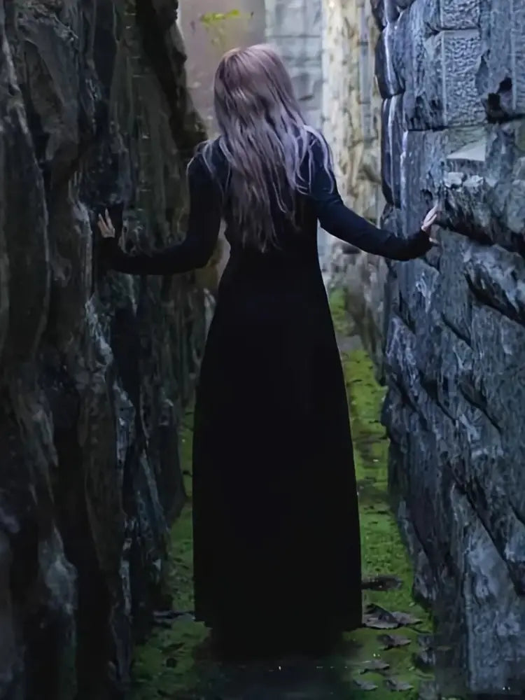 witchy women in a long black dress standing between two stone walls in a dimly lit alley.