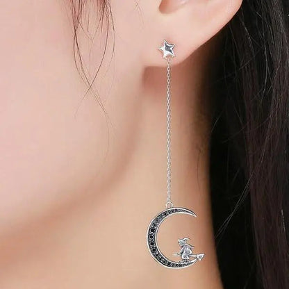 Silver earrings with a moon and witch design worn by a person.