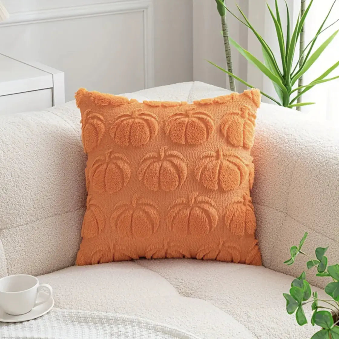 Orange pillow with pumpkin pattern on a beige sofa with a cup and plant in the background