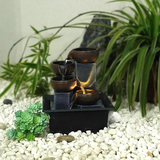 Small water fountain with three bowls on a bed of white pebbles with green plants in the background.