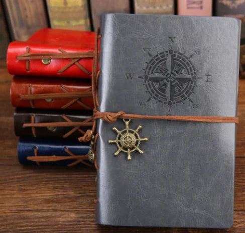 Gray leather notebook with compass design and leather strap, surrounded by other notebooks on a wooden surface.