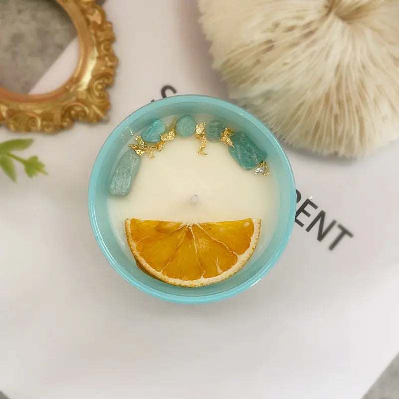 Candle in a teal container with dried orange slice and decorative stones on a textured surface.