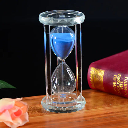 Glass hourglass with blue sand on a wooden surface next to a red book