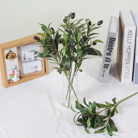 Artificial olive branches in a clear vase on a white surface with books and a photo frame in the background.
