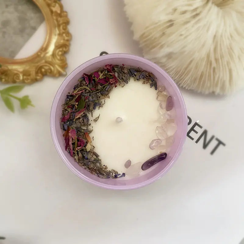 Candle with dried flowers and herbs in a pink container on a white surface.