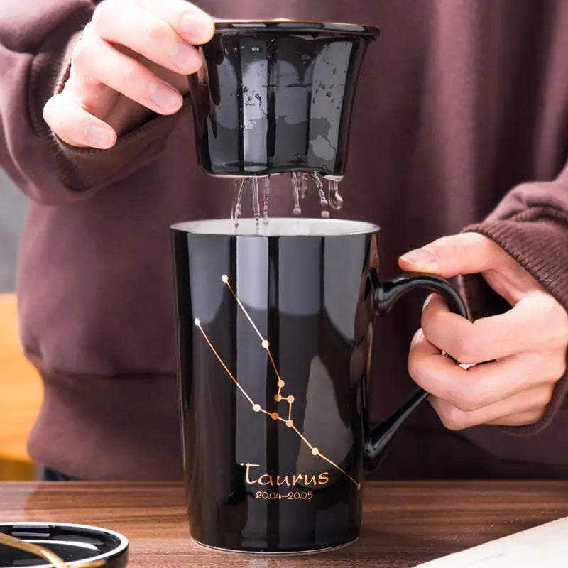 Person pouring liquid into a black mug with 'Taurus' design, held by hands against a blurred background.