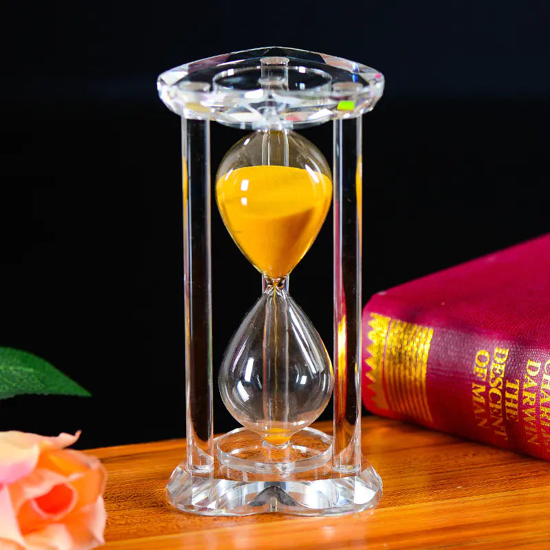 Sand timer with yellow sand on a wooden surface with a book and flower in the background