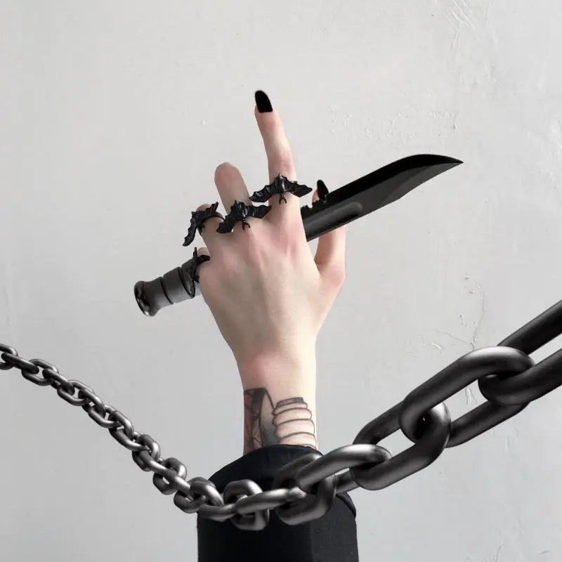 Hand holding a black knife with multiple rings and tattoos, surrounded by chains on a light gray background