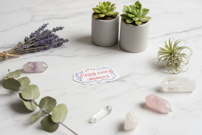 Hand holding a sticker with 'Enjoy every moment' text in front of a colorful circular design.
